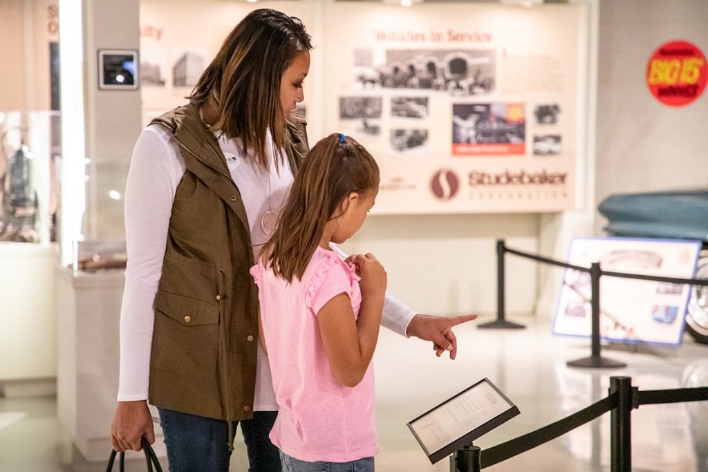 A mom and her daughter read signage in the Museum's Second Floor Gallery. The girl is in a pink short sleeve shirt and the mom is wearing a brown vest over a white long sleeve shirt.