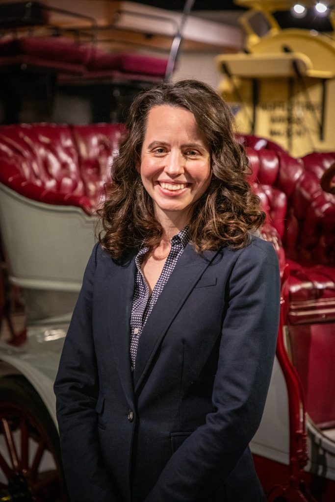 A woman stands in front of a grey car with red upholstery. She is wearing a blue blazer.